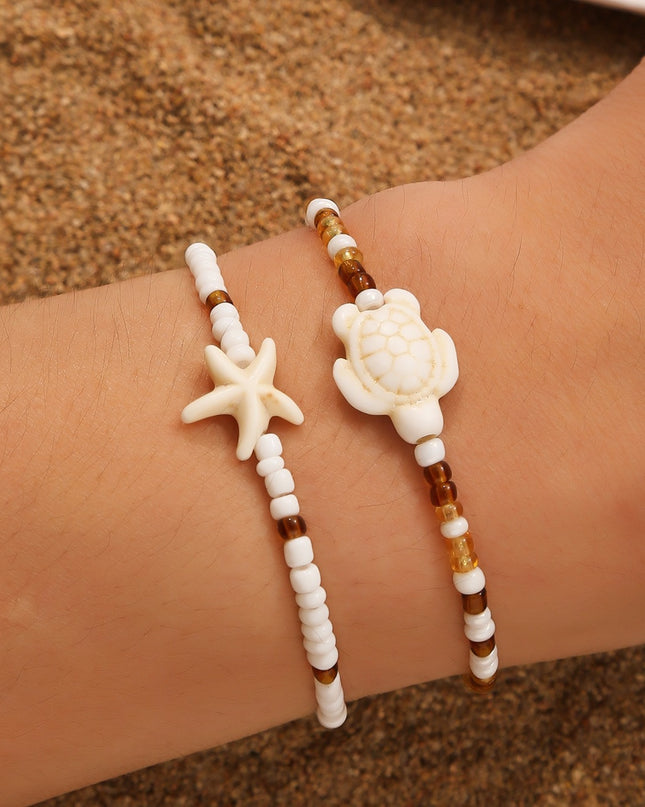 Two beaded bracelets with starfish and turtle designs on a wrist against a sandy background.