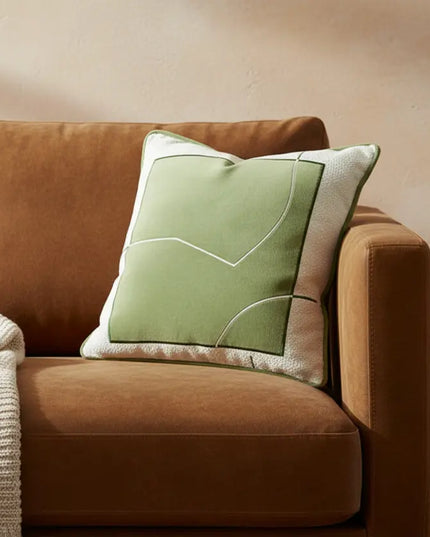 Brown sofa with a green pillow and a blanket, next to a small wooden table with a mug and book.
