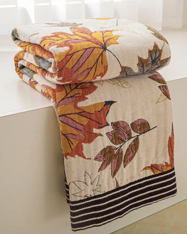 A folded quilted blanket with an autumn leaf pattern in orange, yellow, and brown on a cream background, draped over the edge of a white bathtub.
