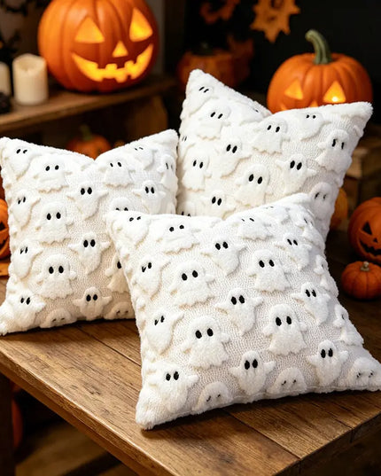 Three white textured throw pillows with a ghost pattern, arranged on a wooden table in a Halloween-themed setting with jack-o'-lanterns and candles.