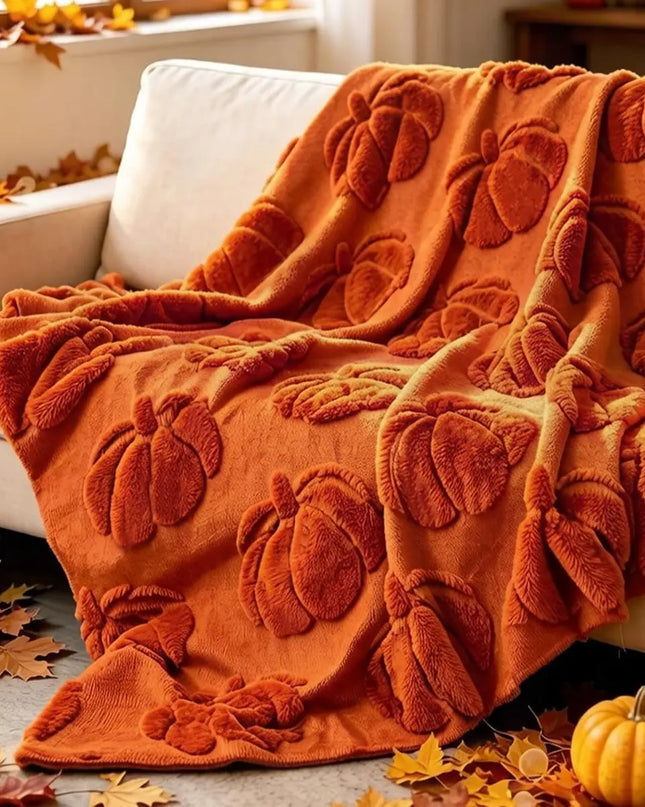 An orange throw blanket with raised pumpkin motifs draped over a white armchair in a cozy indoor setting with autumn leaves and pumpkins on the floor.