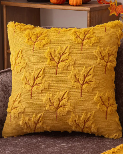 A square yellow throw pillow with a textured knit leaf pattern, resting on a gray upholstered chair in a cozy indoor setting.