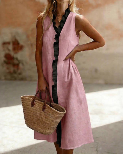 Woman wearing a sleeveless pink and black ruffle-trim linen midi dress, holding a woven straw tote bag, standing in sunlight against a rustic wall