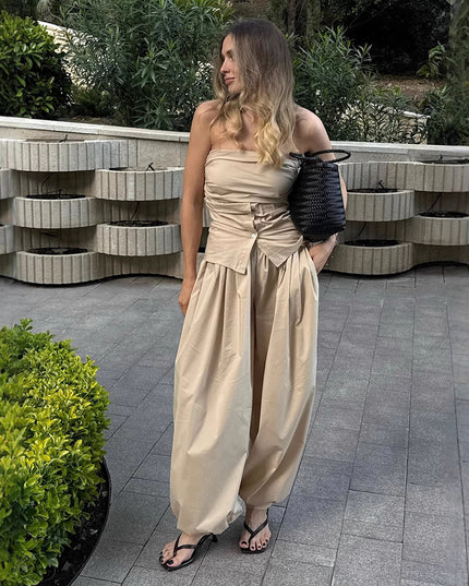 Woman in a beige strapless jumpsuit standing outdoors with greenery and a stone wall in the background.