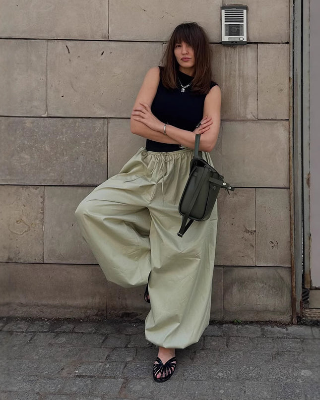 Woman in black top and light green pants leaning against a stone wall.