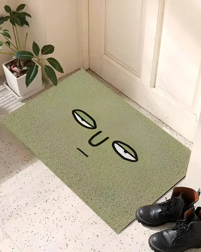 Green doormat with a face design on a tiled floor next to a plant and shoes.