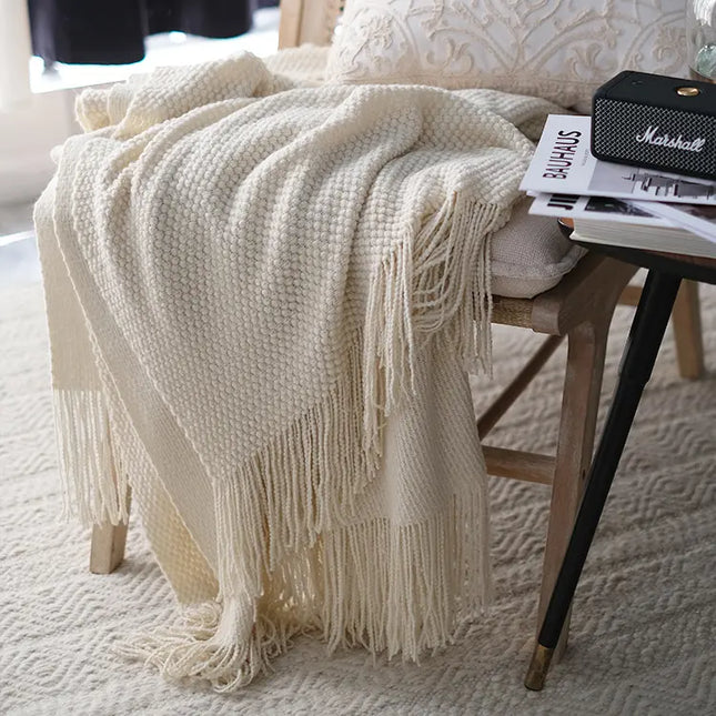 Beige knitted throw blanket with fringe draped over a chair in a cozy room.