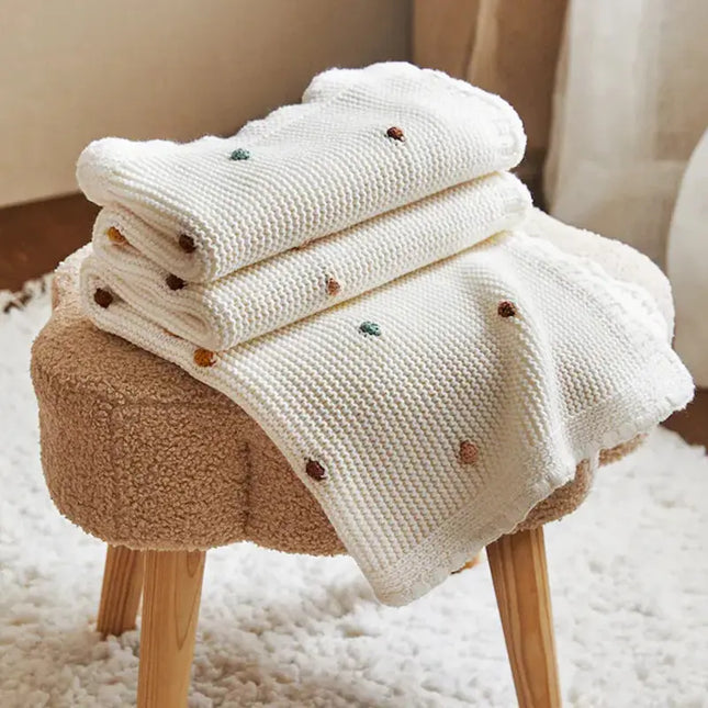Stack of white baby blankets with embroidered dots on a wooden stool.