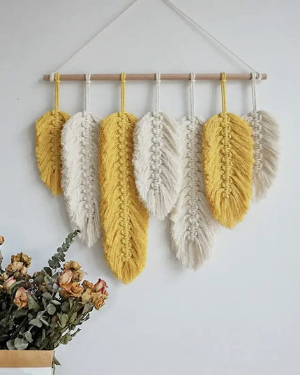 Hanging macrame wall art with yellow and white feathers on a plain wall.