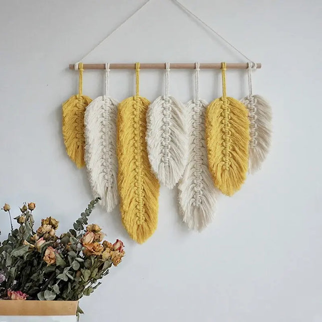 Hanging macrame wall art with yellow and white feathers on a plain wall.