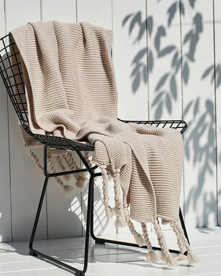 Beige knitted blanket with tassels draped over a black metal chair against a white wooden wall.