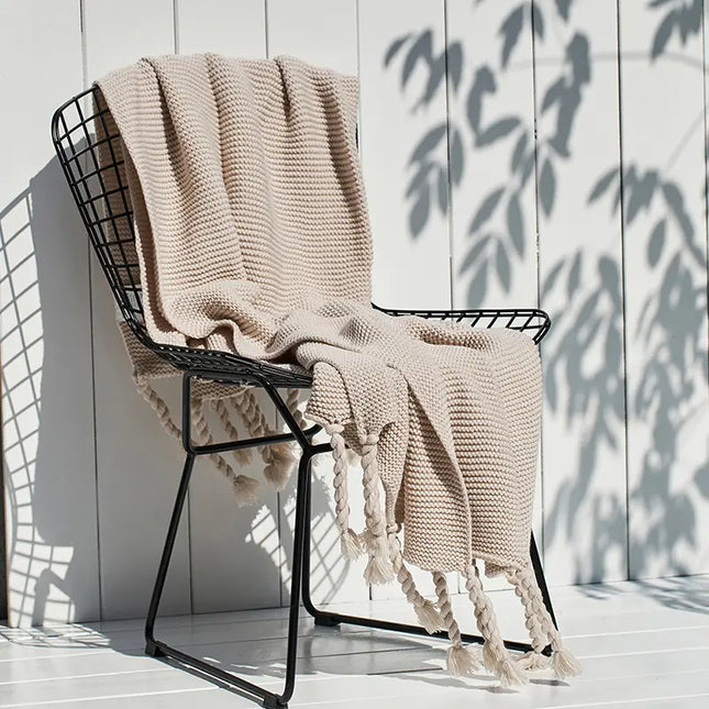 Beige knitted blanket with tassels draped over a black metal chair against a white wooden wall.