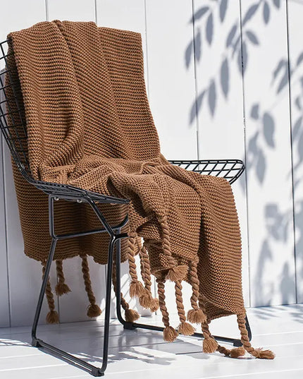 Brown knitted blanket draped over a black metal chair against a white wooden wall.