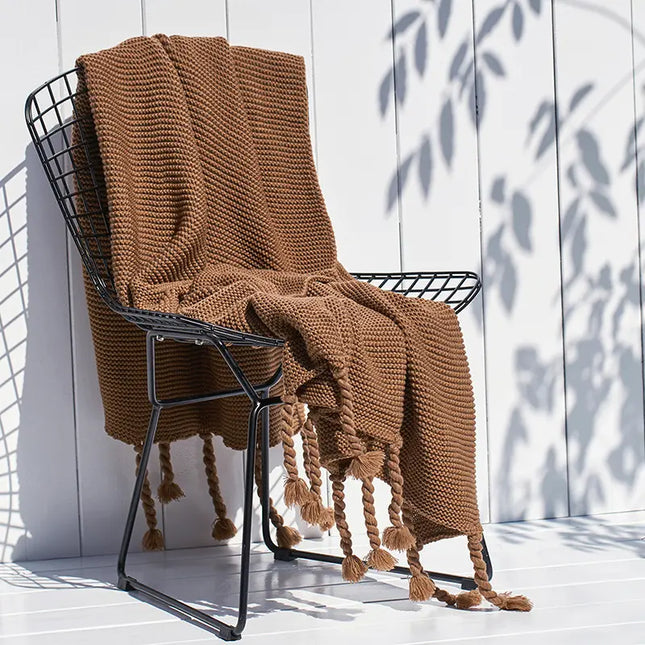 Brown knitted blanket draped over a black metal chair against a white wooden wall.