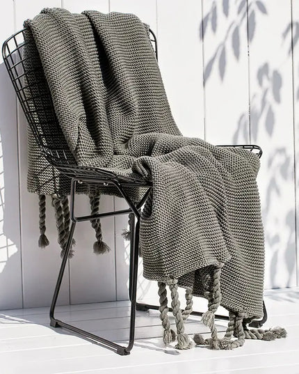 Gray knitted blanket draped over a black metal chair on a white wooden floor with a white wall in the background.