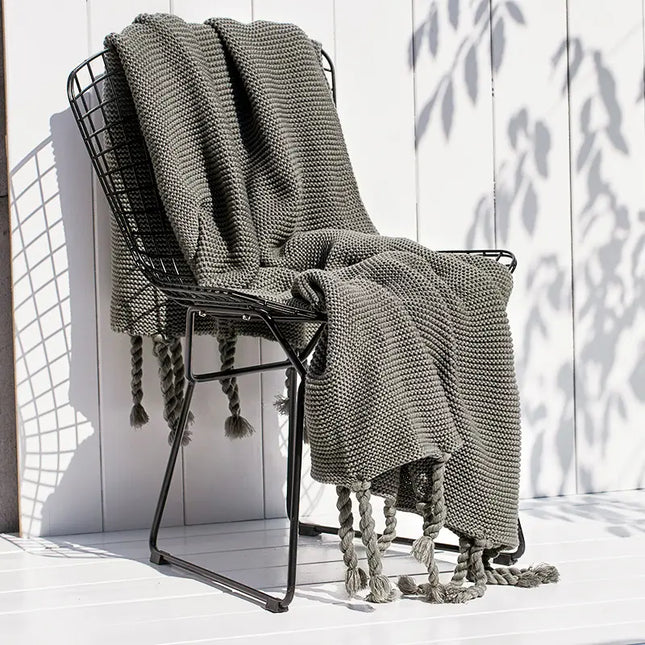 Gray knitted blanket draped over a black metal chair on a white wooden floor with a white wall in the background.