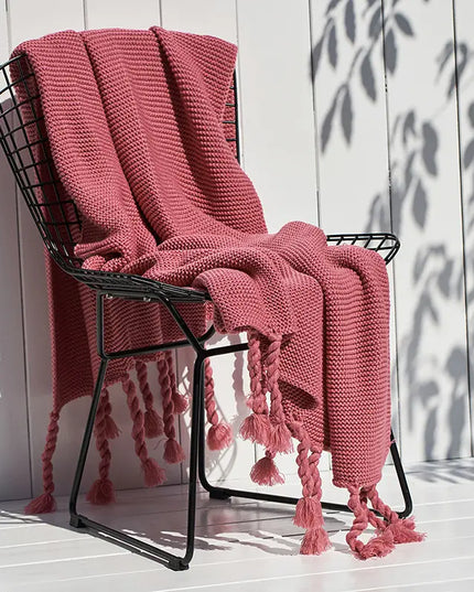 Pink knitted blanket with tassels draped over a black metal chair against a white wooden wall.