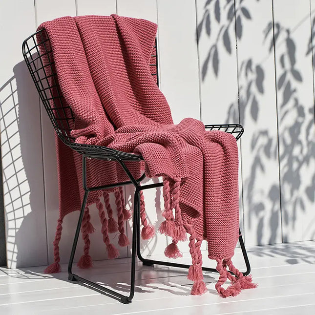 Pink knitted blanket with tassels draped over a black metal chair against a white wooden wall.